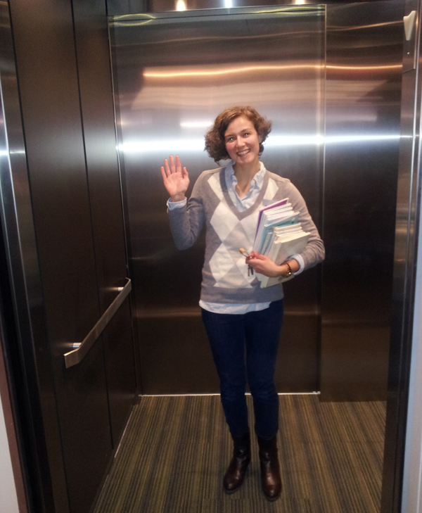 Librarian holding a stack of books smiles and waves from inside the new elevator