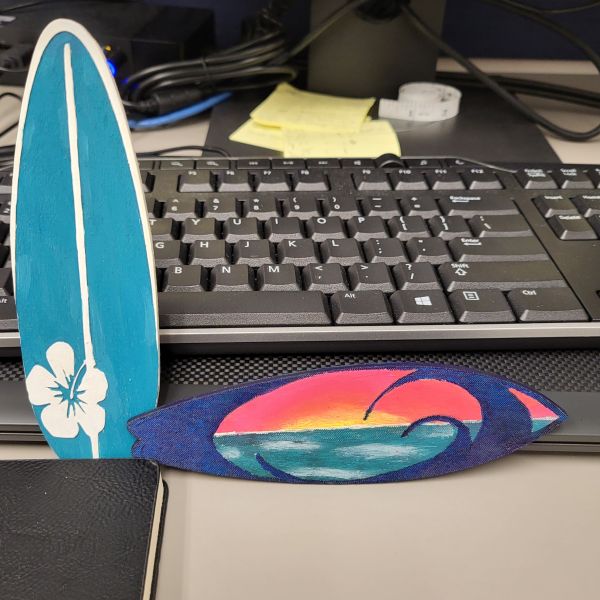 Two mini surfboards etched and cut on the laser. One painted in blue and white with a hibiscus flower, and the other with a blue swirl and the sunsetting over the ocean on the other.