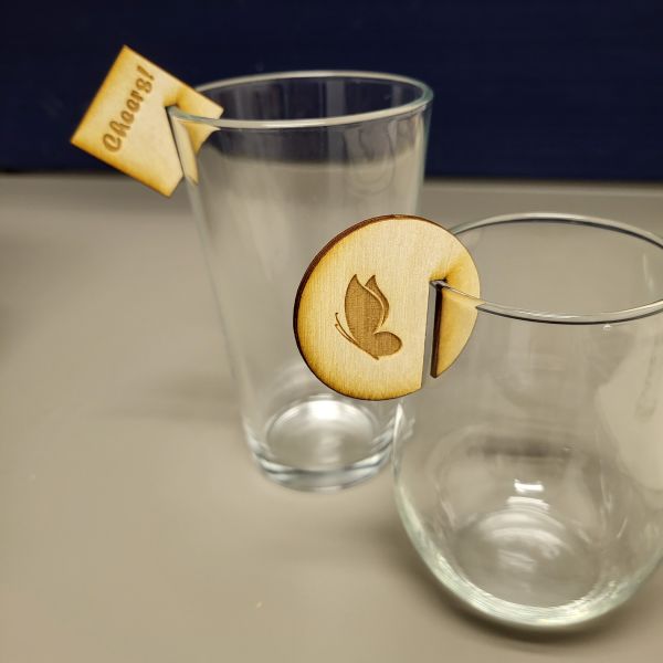 A square drink marker that says Cheers! on it hangs on the rim of a pint glass. Round drink marker with a butterfly silhouette on a stemless wine glass.