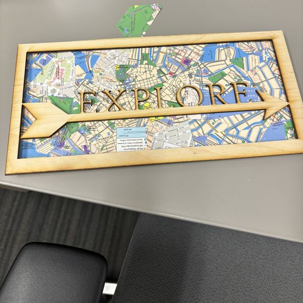 The word EXPLORE on an arrow shape and cut out with a frame on birch wood. Back panel cut out of birch wood and covered with collaged map pieces modpodged on.
